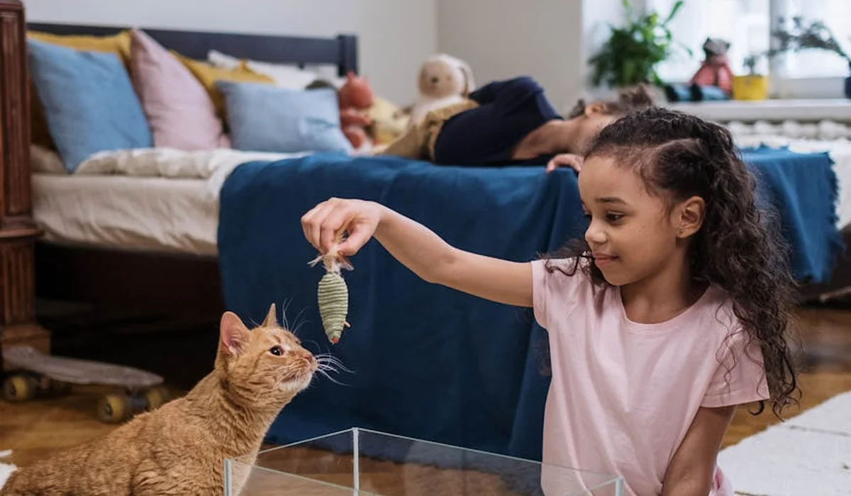 A young girl holds a fish above a clear fish tank while a curious cat watches from the side; a bedroom with a bed, pillows, and stuffed animals is visible in the background.