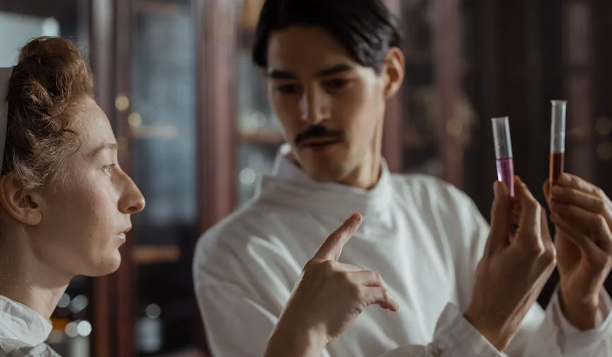 Two researchers in lab coats discuss ammonia testing, one gesturing toward two test tubes held by the other, in a laboratory setting.