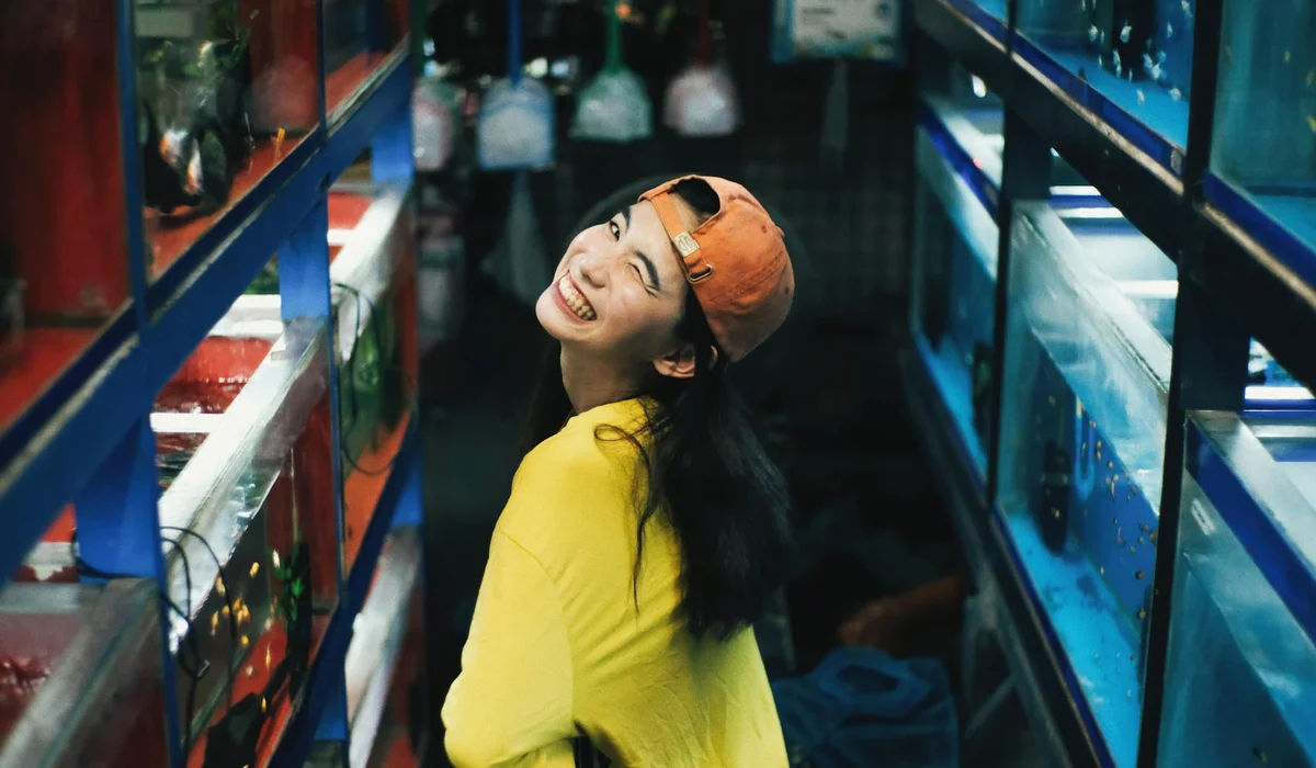 Smiling shopper in a yellow shirt and orange cap browses aquarium supplies in a store aisle with tanks on both sides.