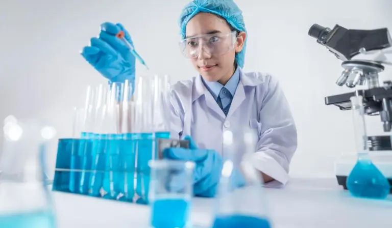 Scientist in safety goggles and gloves uses a pipette to transfer blue liquid in a lab with test tubes and a microscope, illustrating lab setup for water testing.