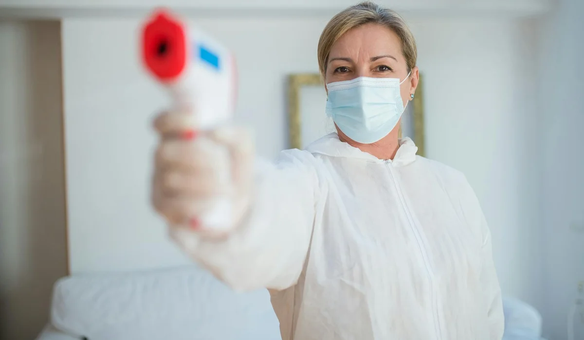 Person in protective PPE and face mask holding a digital thermometer toward the camera.