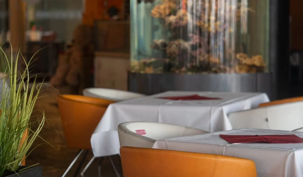 Dining area with orange chairs, white-tablecloth tables, and a large aquarium in the background.