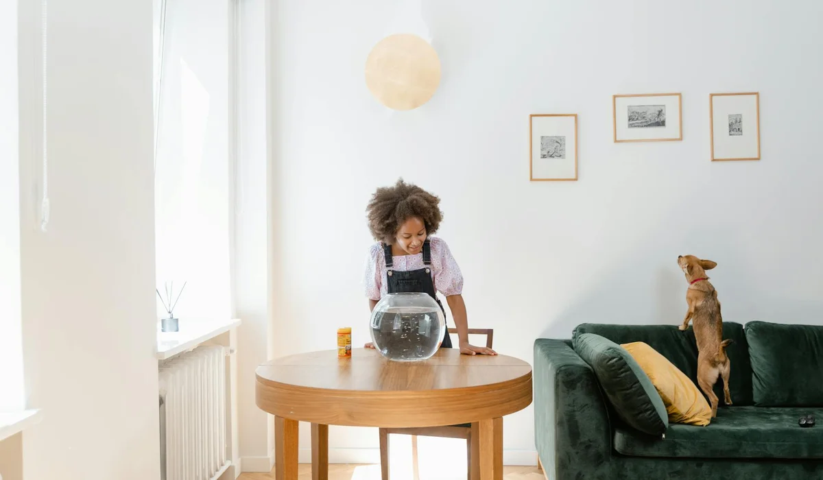 A bright living room with a woman standing at a round wooden table, looking into a small glass fish bowl; a dog sits upright on a green sofa to the right, and sunlight streams in from a window on the left.