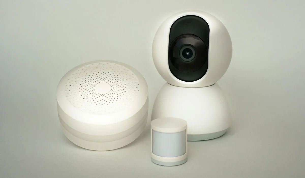 White security camera, a motion sensor, and a small smart hub on a neutral background