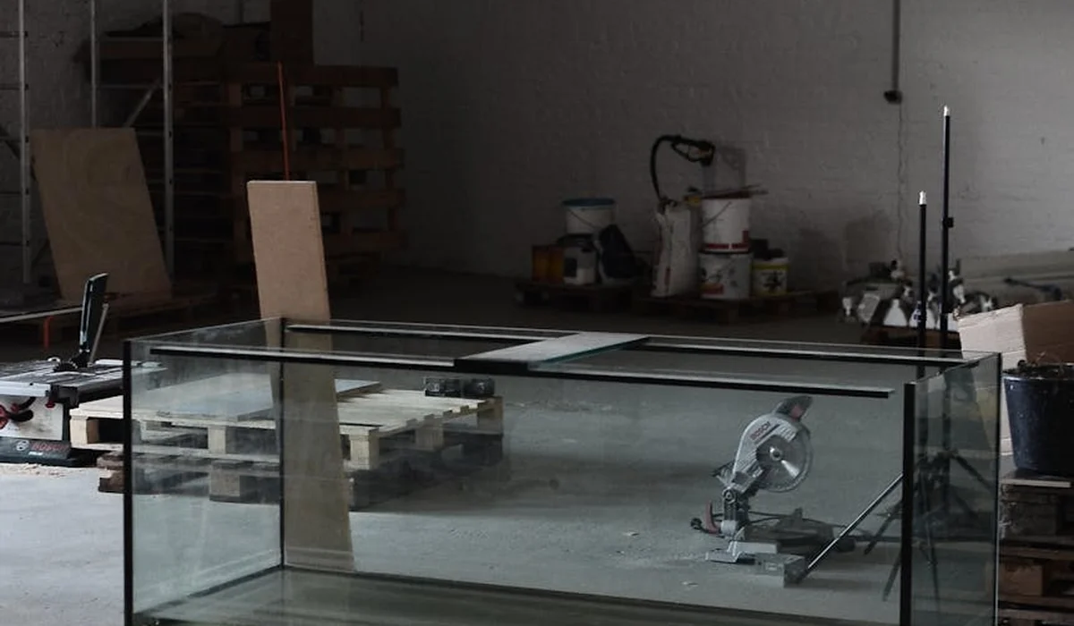 A glass aquarium sits on a stand in a dim, cluttered workshop with tools and supplies in the background.