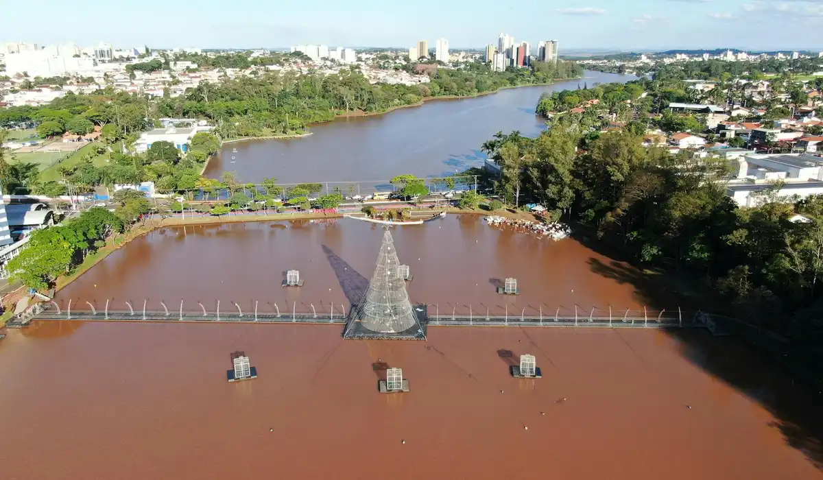 Aerial view of a tea-colored blackwater pond with a central promenaded structure, surrounded by trees and an urban skyline in the distance.