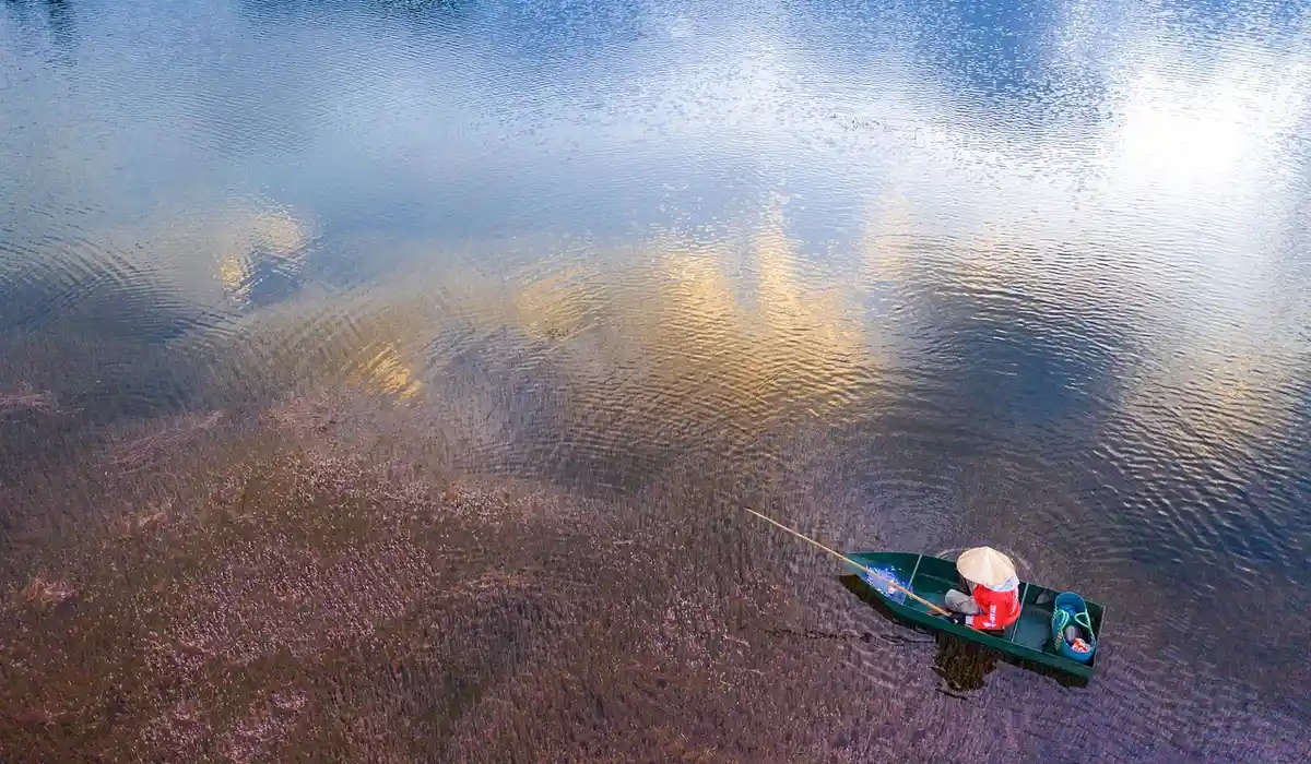 A small boat with a person wearing a white hat sits on a calm lake, with clear water and reflections on the surface.