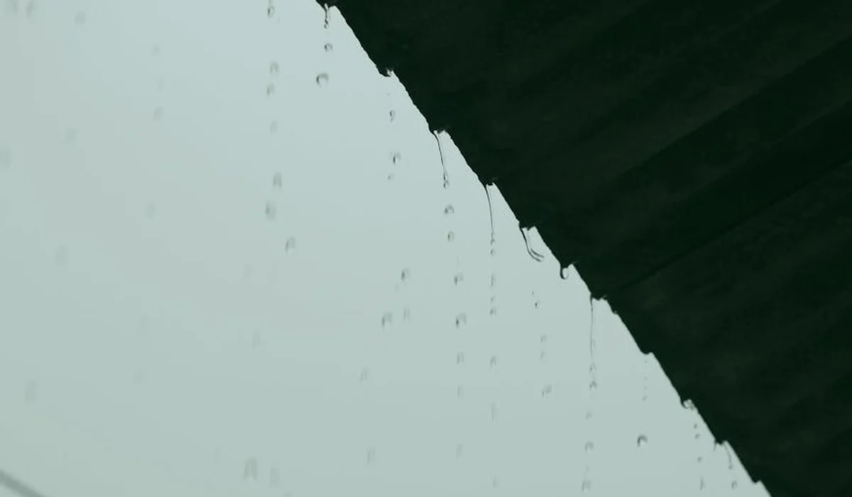 Water droplets dripping from the edge of a roof against a pale sky.