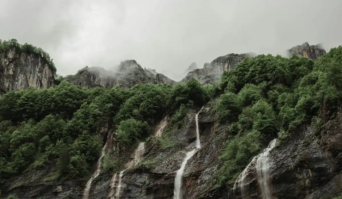 Lush green forest with rocky cliffs and multiple small waterfalls cascading down, under a cloudy sky.