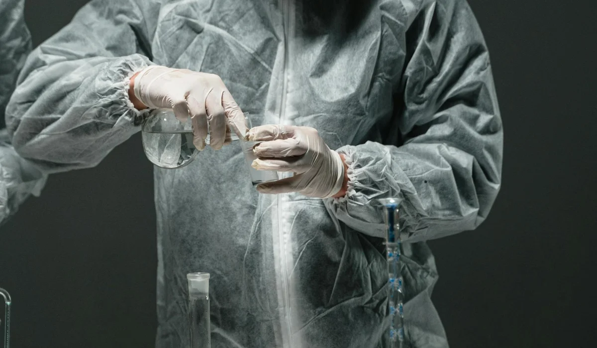 A researcher in a protective lab gown pours liquids and prepares glassware, illustrating careful handling while reading aquarium test strip colors.