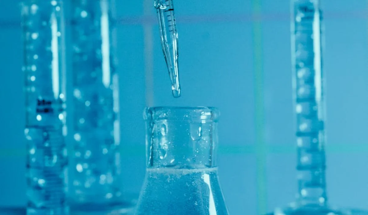 Close-up of a dropper releasing liquid into a glass beaker in a blue-tinted laboratory setting, illustrating water quality testing for aquariums.
