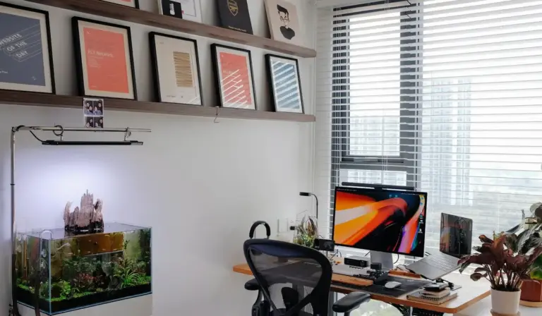 Home office with a planted aquarium on the left, a computer desk on the right, framed certificates on the wall, and a window with blinds.
