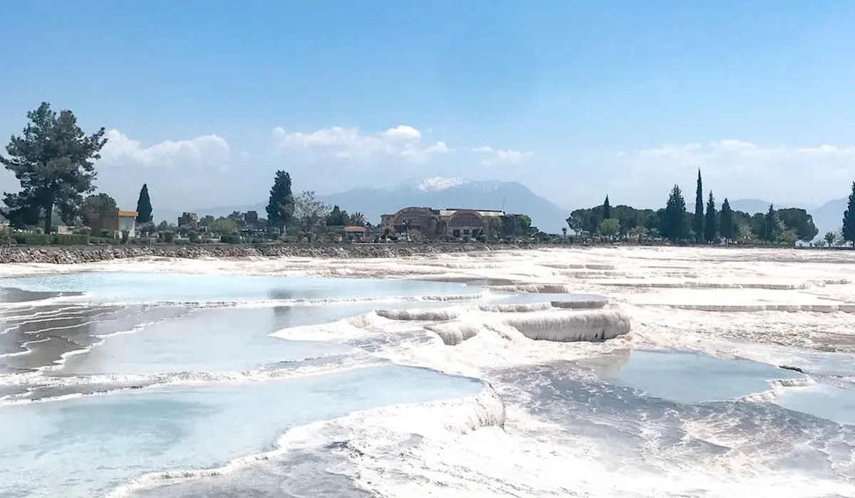 White travertine terraces with shallow blue pools under a clear sky, with distant buildings and trees along the horizon.