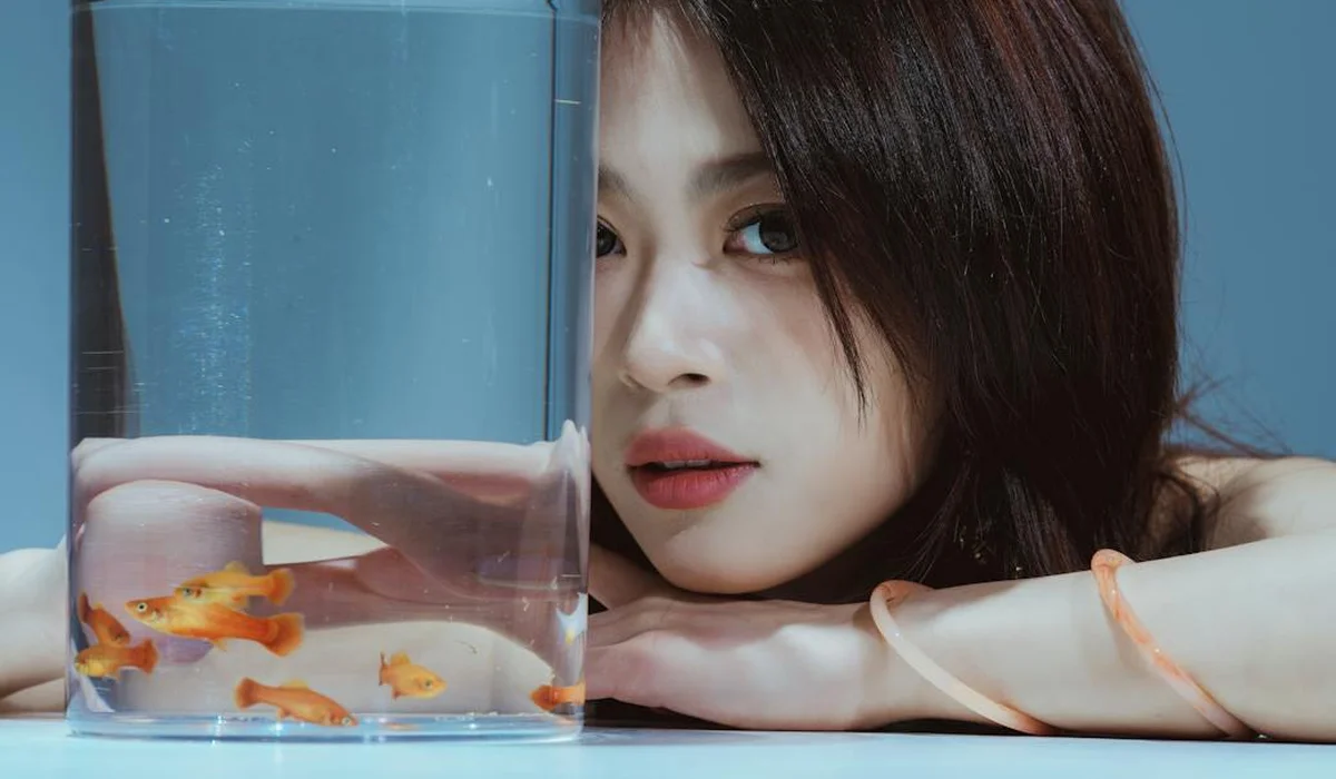 Close-up of a person leaning on a glass aquarium filled with small orange fish, resting on a blue surface