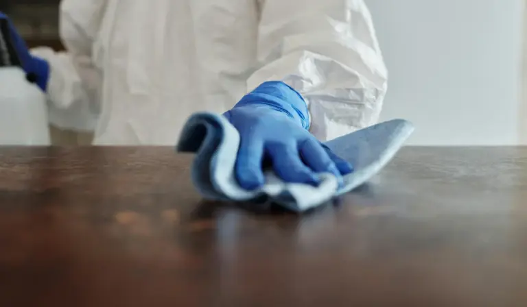 Person wearing white protective gear and blue gloves wipes a wooden surface with a blue cloth