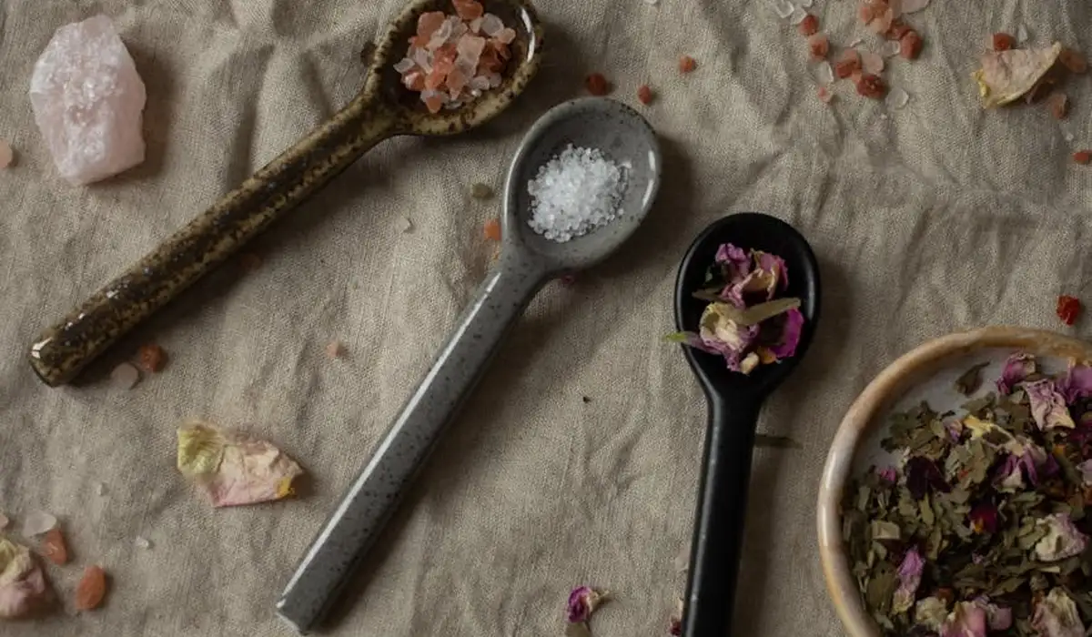 Close-up of three measuring spoons filled with salt crystals and pink Himalayan salt on a beige fabric, with a small bowl of dried flower petals nearby.