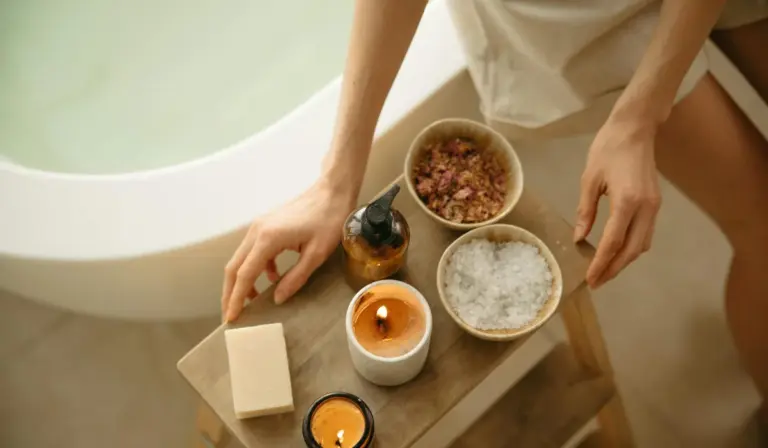 Top-down view of a wooden tray placed beside a bathtub, holding bath salts, a bar of soap, a small bottle, and lit candles.