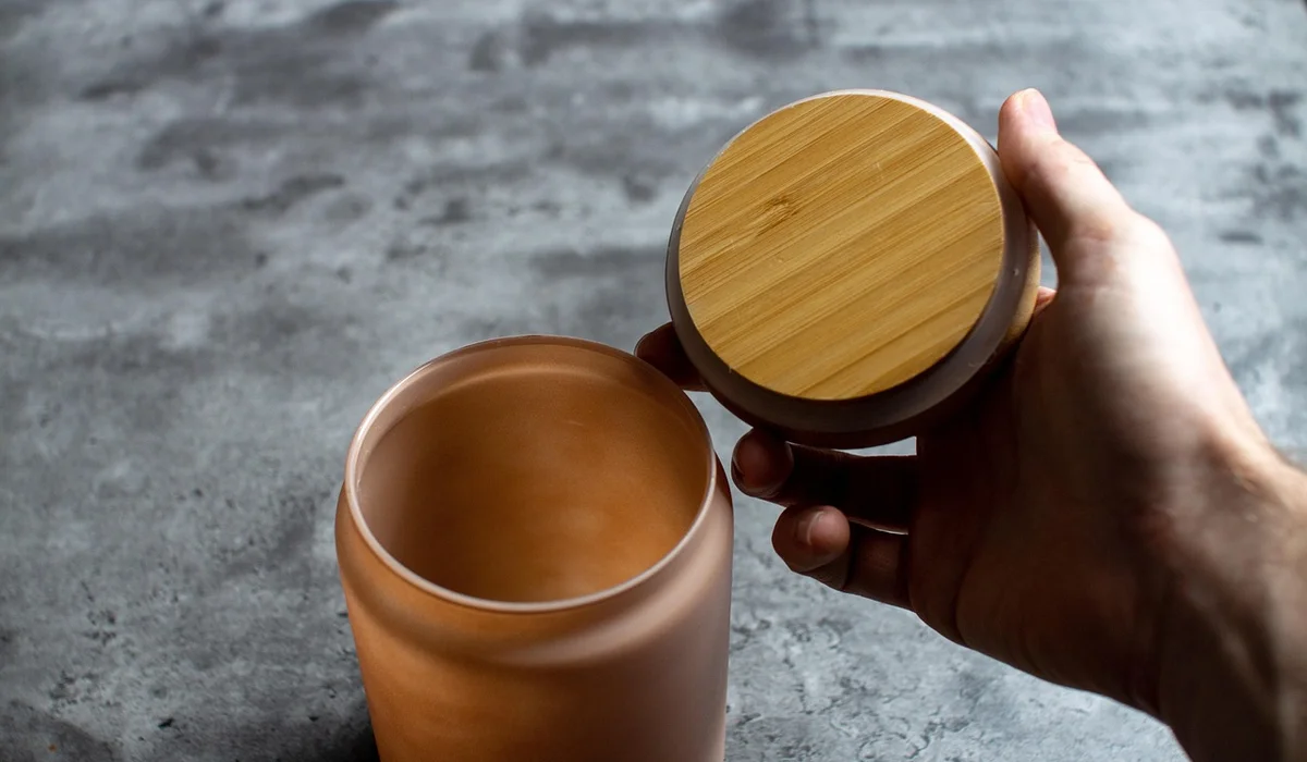 A hand holds a round lid with a bamboo top and a gray rim beside an open cylindrical container on a concrete surface, illustrating a DIY lid concept for non-standard aquarium tanks.