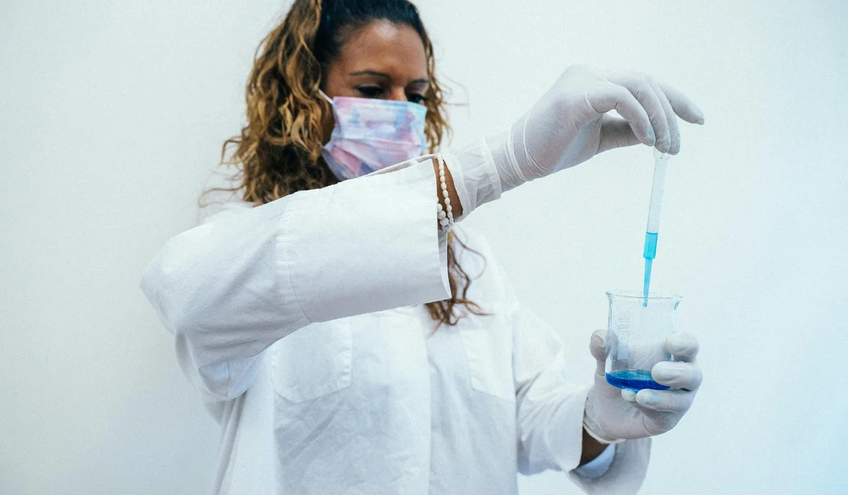 A person in a white lab coat, gloves, and a mask uses a pipette to transfer blue liquid into a small cup.
