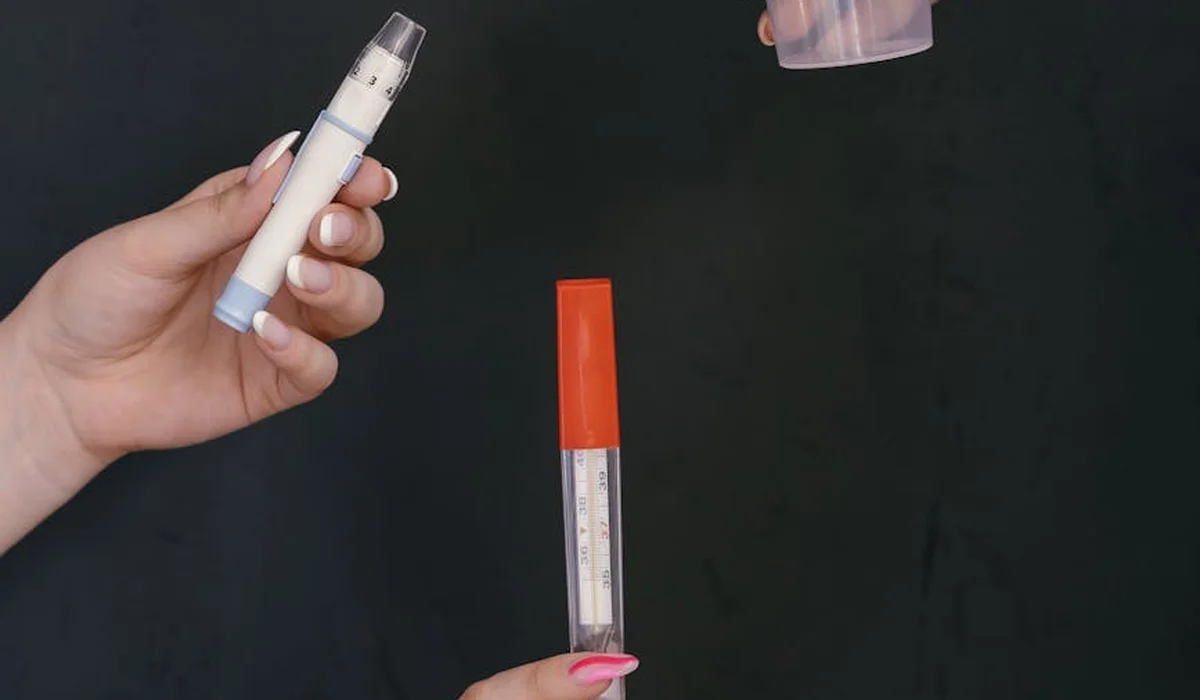 Close-up of hands holding water testing instruments, including a reagent vial in one hand and a red-topped test device with measurement scale in the other, set against a dark background.
