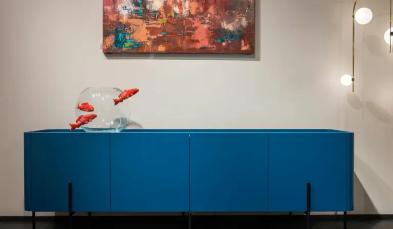 Blue cabinet with a glass fish bowl containing orange fish on top, with a painting on the wall and round lights in the background.