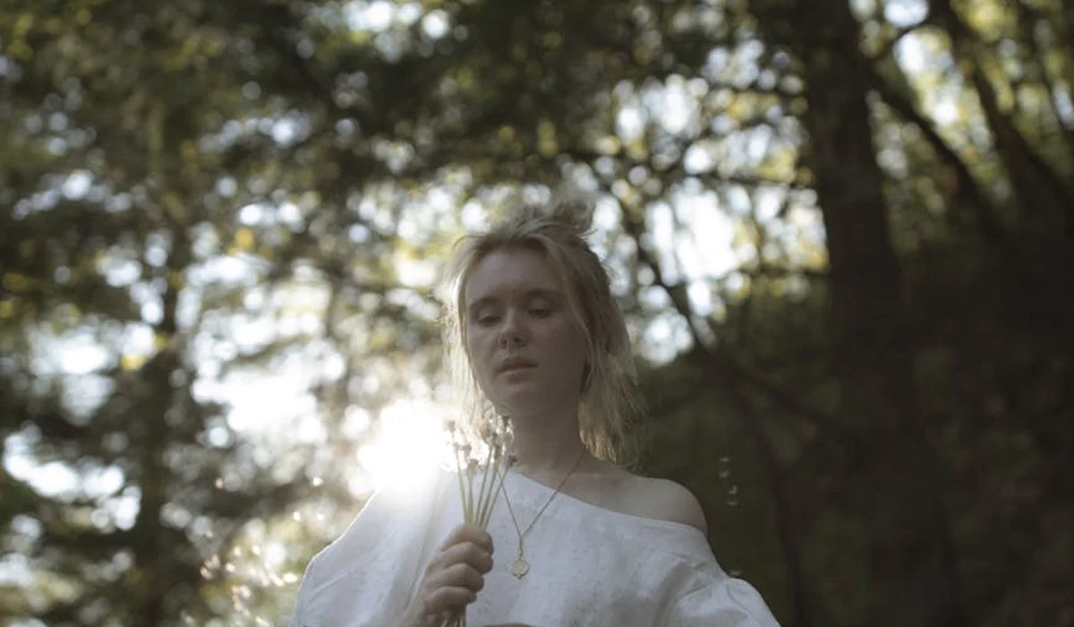 Woman with blond hair standing in a sunlit forest, wearing a white top, symbolizing calm preparation for emergencies.