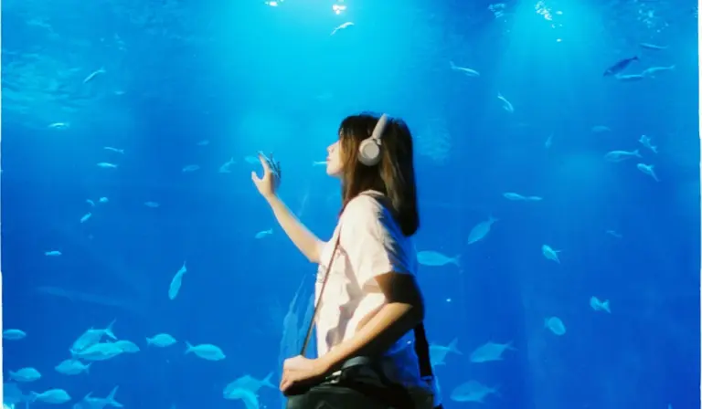 Person wearing headphones standing beside a large blue aquarium filled with swimming fish, seen from the side.