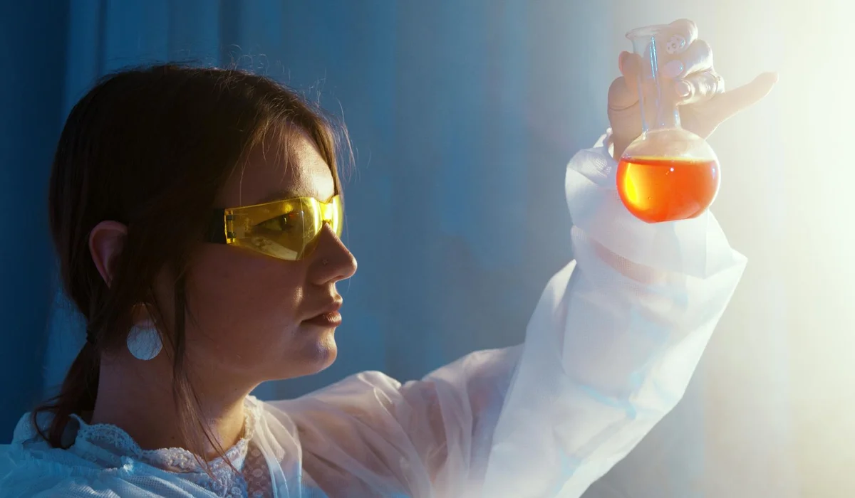 A person wearing safety goggles and a lab coat holds a flask with an orange liquid, examining a water sample for aquarium cycling.