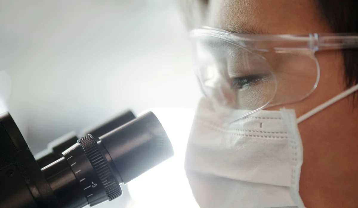 Close-up of a lab technician wearing a face mask and safety goggles, looking into a microscope.