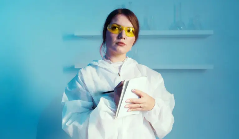 A lab worker wearing a white protective coat and yellow safety goggles holds a notebook in a blue-toned laboratory.