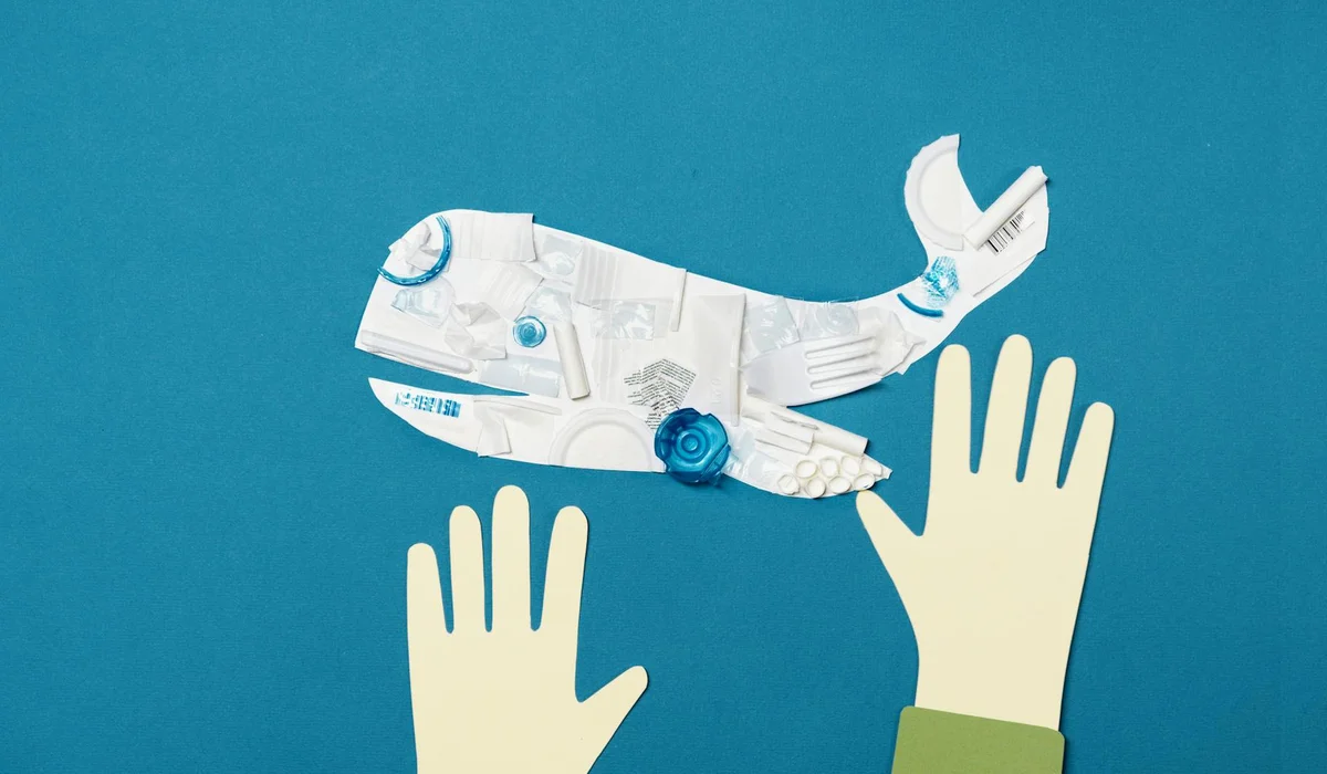 Two pale hands reaching toward a white paper whale model on a blue background.