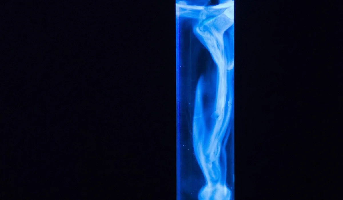 Vertical glass test tube glowing blue with wispy white vapor against a dark background.