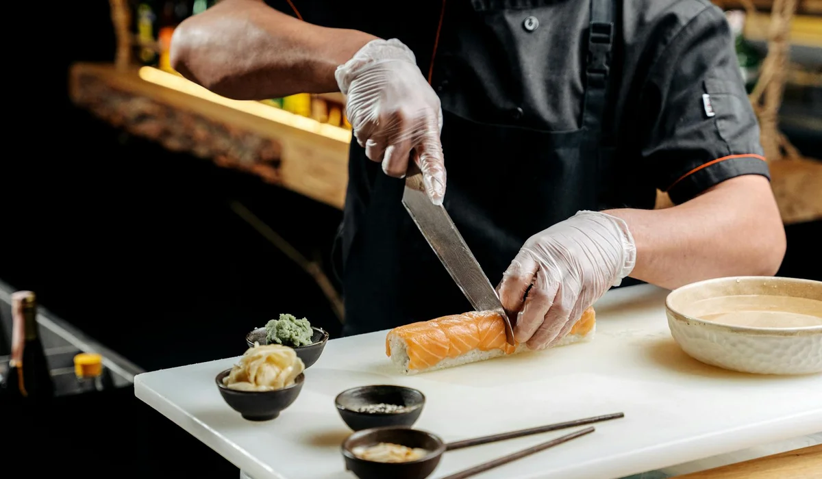 Chef in a dark uniform wearing gloves slices sushi on a cutting board, with small bowls of sauce nearby.