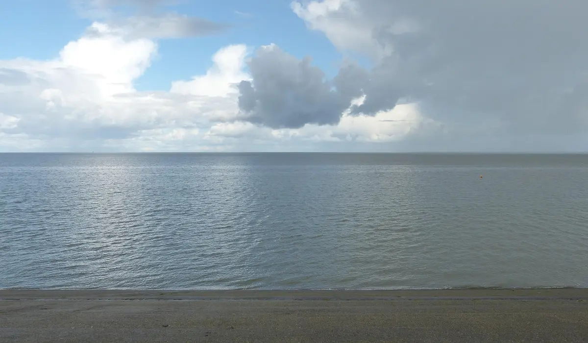 Calm ocean shoreline with the horizon, clouds, and a sandy beach.