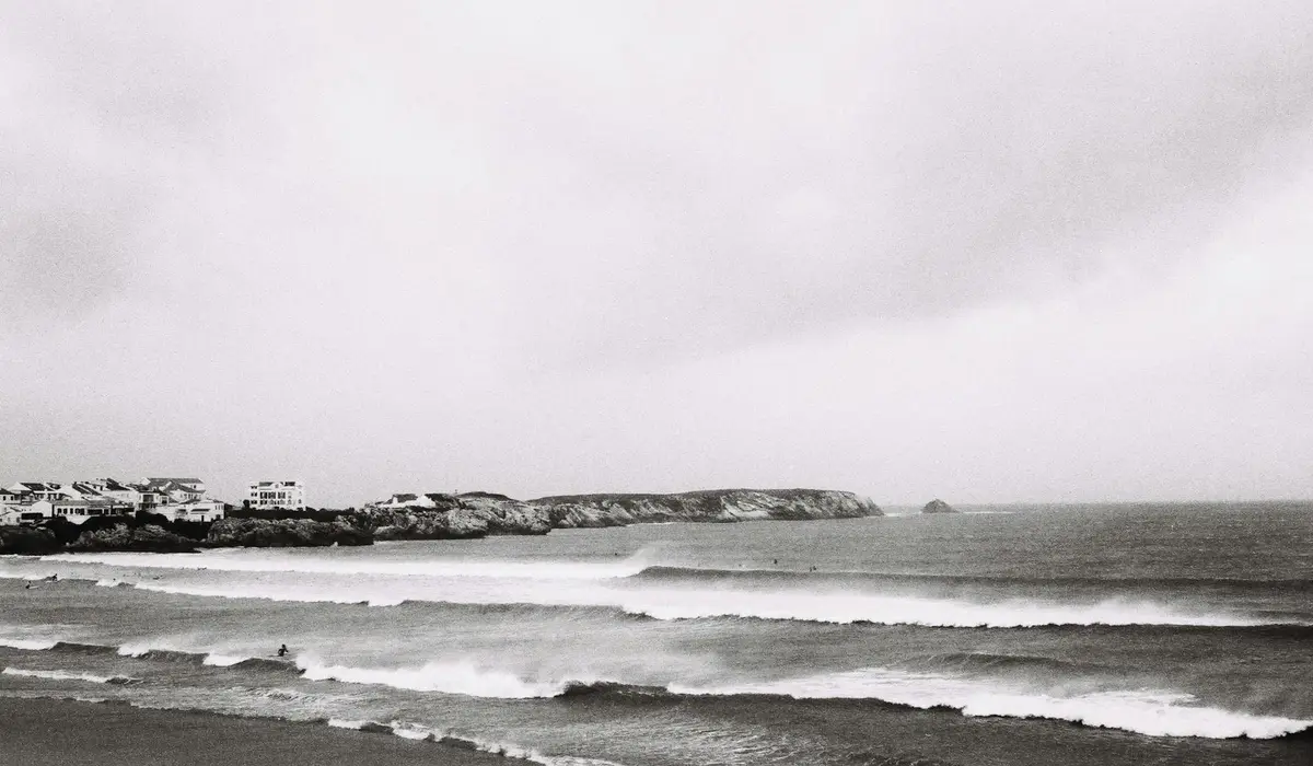 Black-and-white coastal scene with waves rolling onto the shore and houses along the left coastline.
