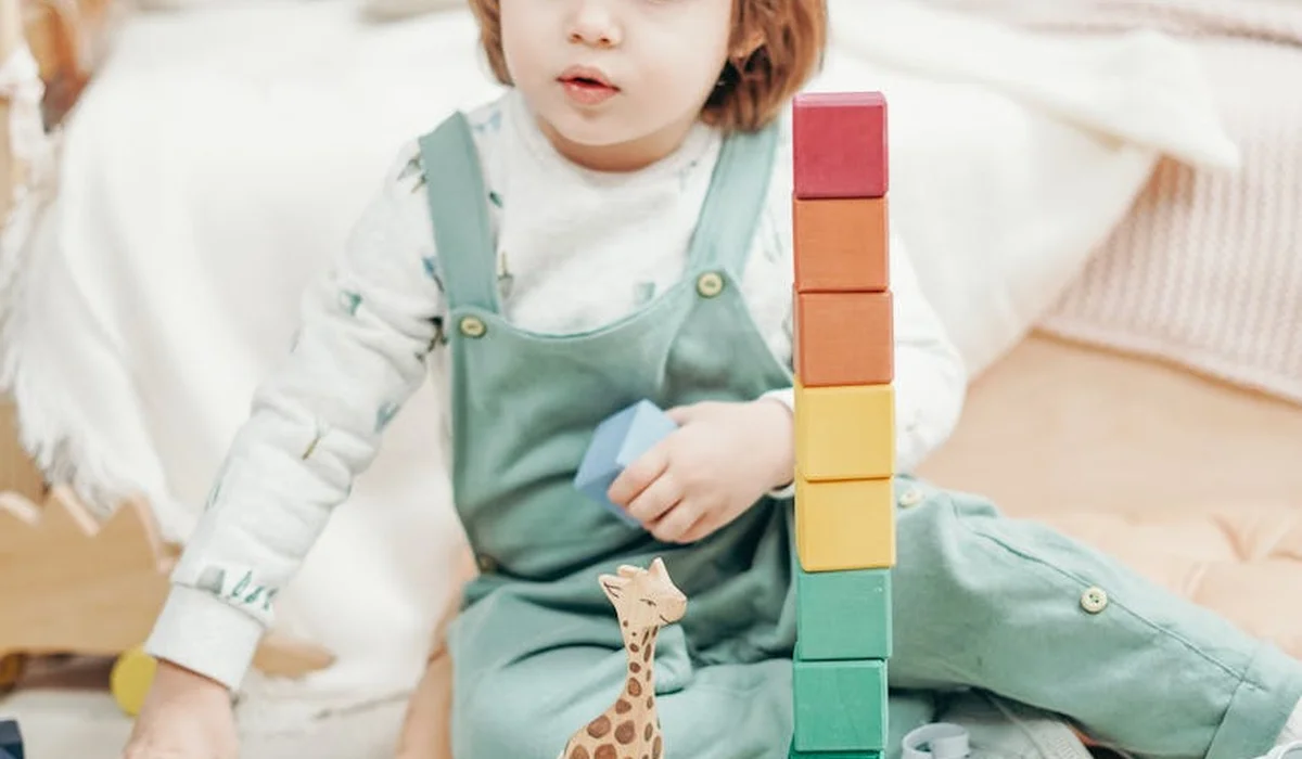 Young child sitting indoors, stacking colorful building blocks with a small toy giraffe nearby.