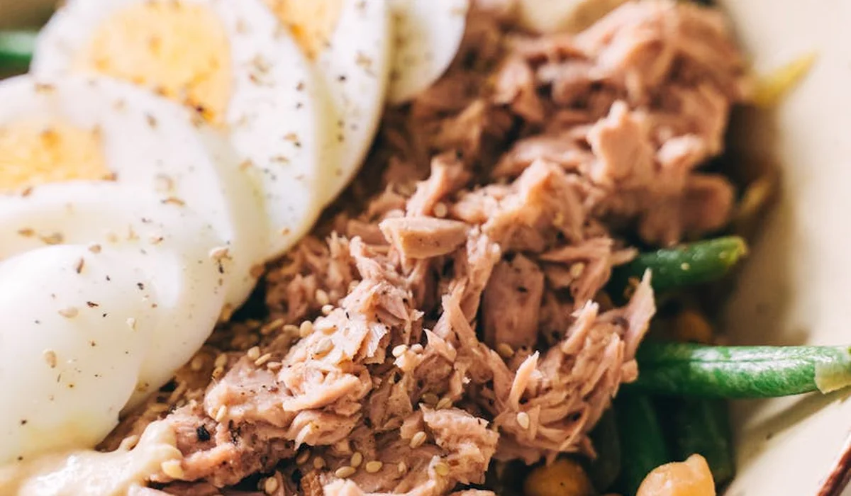 Plate with flaked tuna and sliced hard-boiled eggs, with greens and potatoes in the background