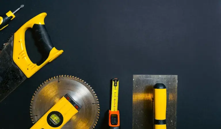 A collection of yellow and black measuring and cutting tools arranged on a dark surface, including a hand saw, a circular saw blade, a tape measure, a square, and a level.