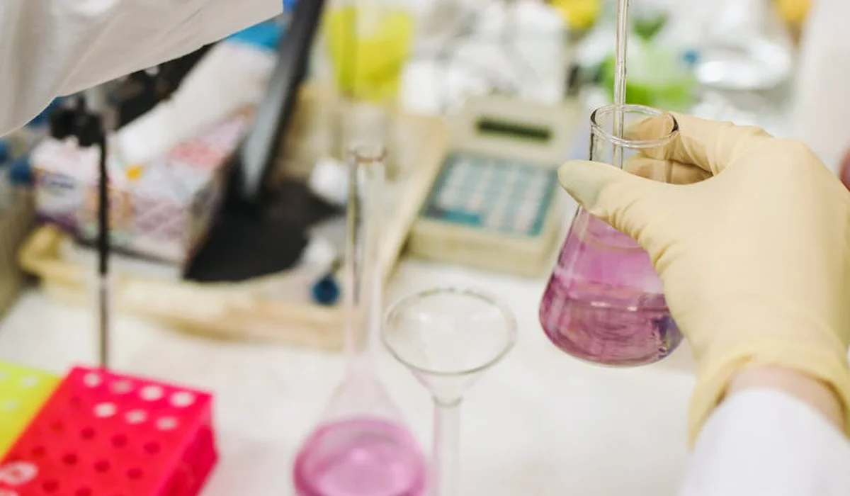 Laboratory scene with gloved hands pouring pink liquid from a flask; various glassware and lab equipment visible in the background, suggesting aquarium water testing.