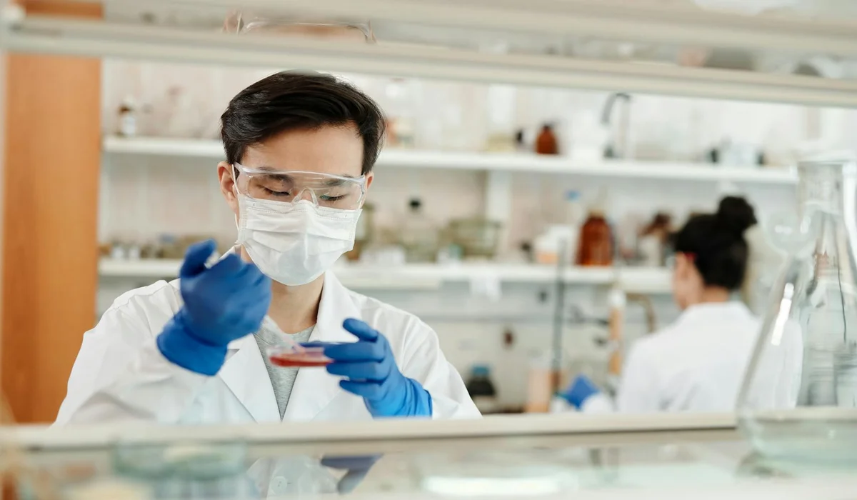 A lab technician wearing safety goggles, a mask, and blue gloves tests a sample at a laboratory bench, illustrating water hardness testing for aquariums.