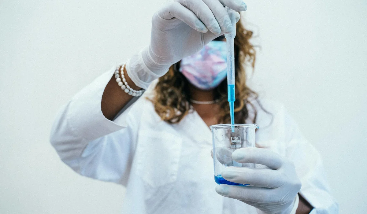 A laboratory scientist wearing gloves and a mask uses a pipette to transfer blue liquid into a beaker.