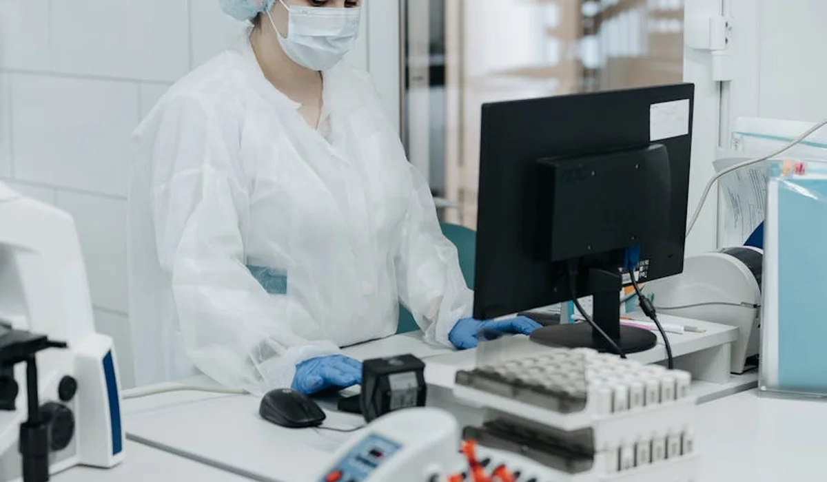 A laboratory technician wearing a mask, gloves, and a white lab coat sits at a bench with testing equipment and a computer, examining water samples.