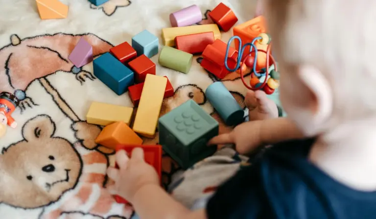A child’s hands playing with colorful LEGO bricks on a soft patterned blanket, with scattered blocks and a red toy accessory in the background.