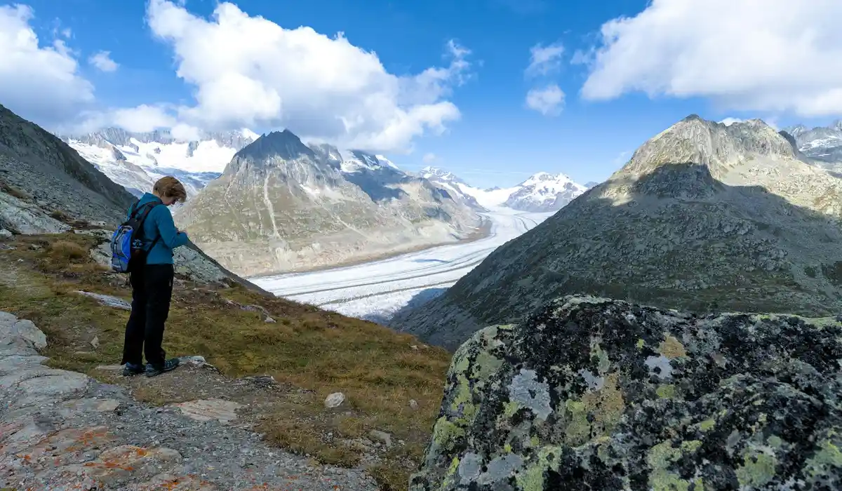 Hiker in a blue jacket standing on a rocky alpine overlook, with snow-capped mountains and a glacier in the distance.