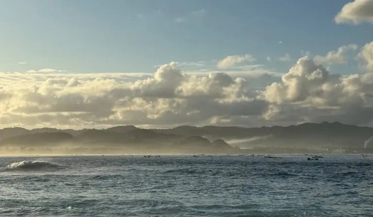 Calm ocean waves at a distant shoreline under a cloudy sky.