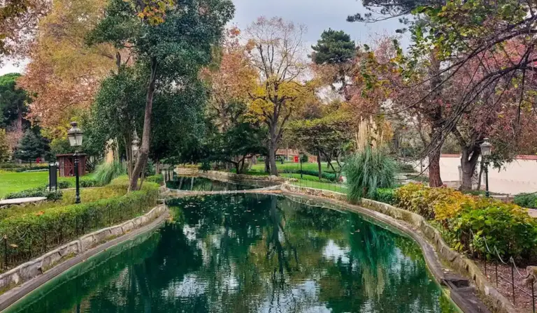 Autumn park scene with a winding, reflective pond surrounded by trees and greenery.