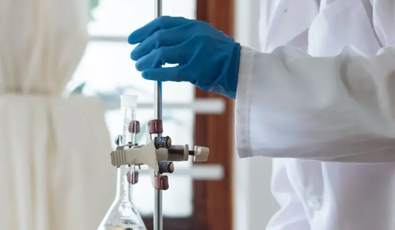 Lab technician in a white coat wearing blue gloves adjusts glassware with clamps in a laboratory setting.