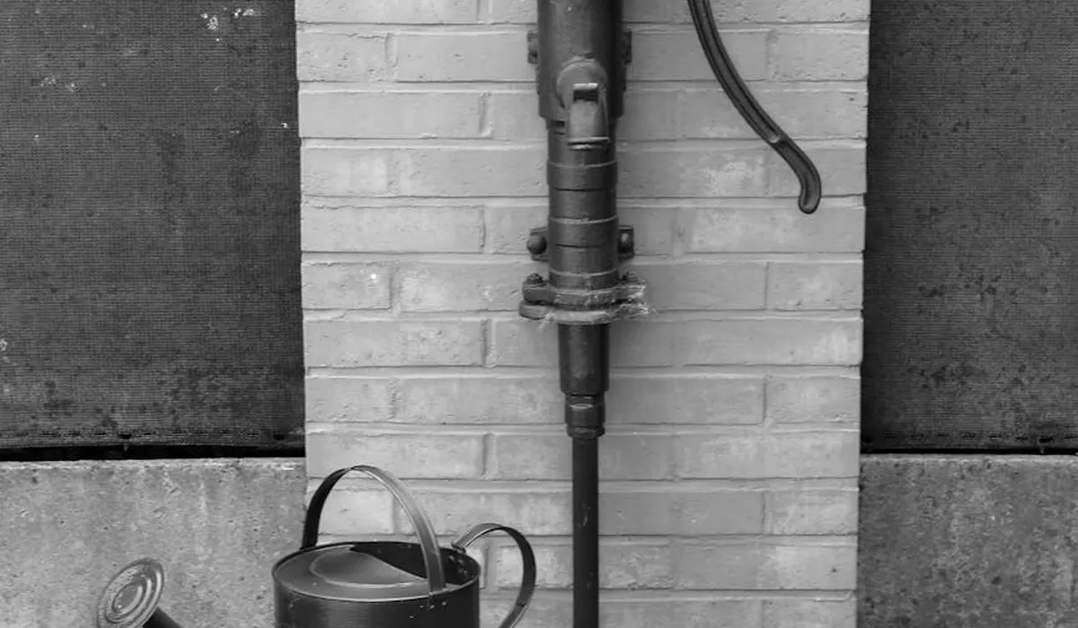 Black-and-white photo of an outdoor water pump mounted on a brick wall, with piping and a watering can nearby.
