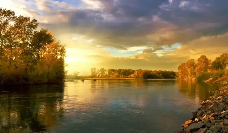 Autumn river scene at sunset with trees in fall colors reflected on calm water.