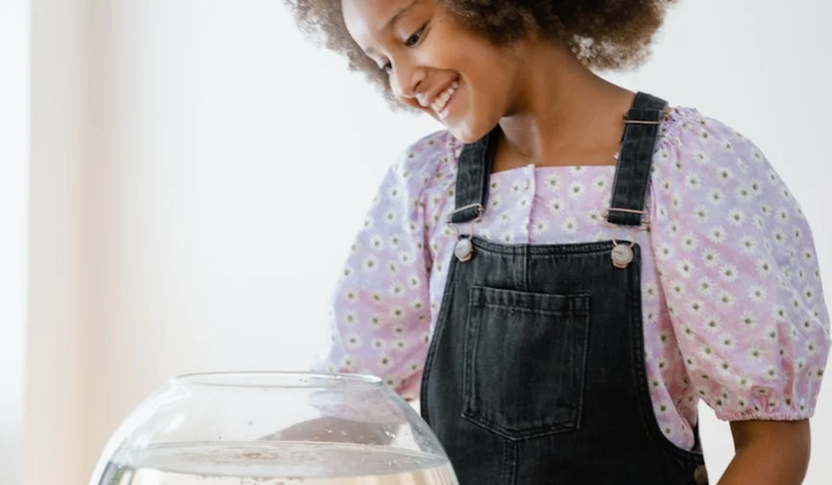 A smiling person wearing a pink polka-dot shirt and dark overalls looks toward a small glass fish tank